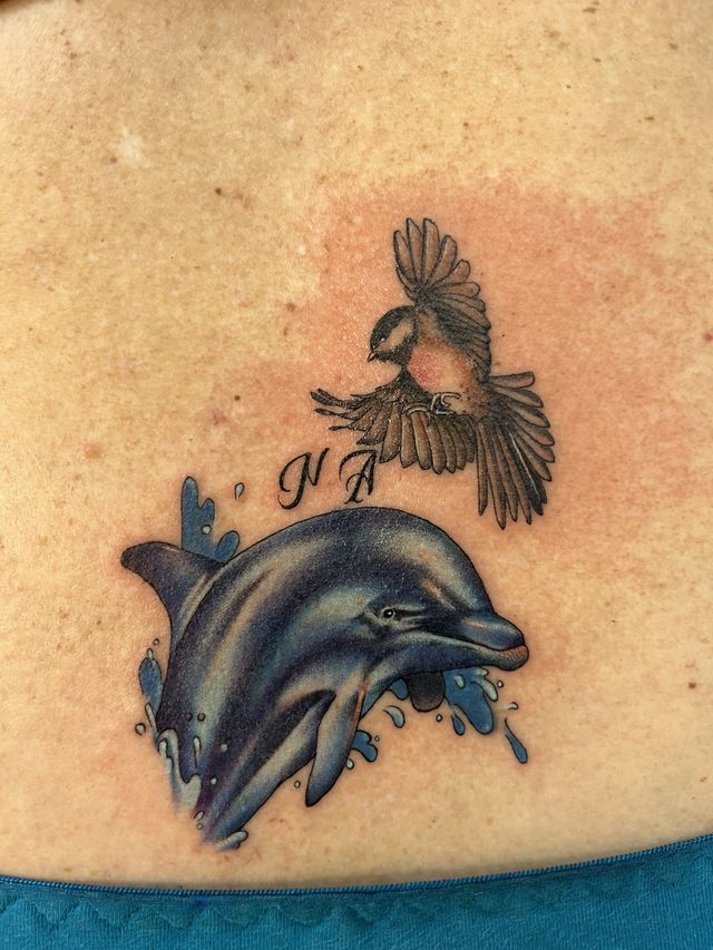 Colour tattoo of a dolphin leaping through blue splashes with a small bird above and two initials.