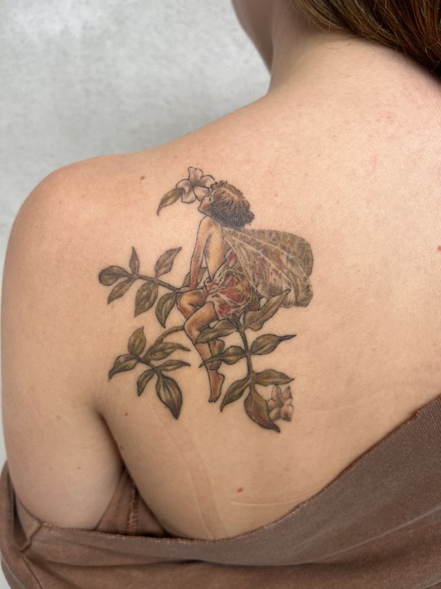 Colour tattoo of a flower fairy sitting in leafy branches on a shoulder blade.